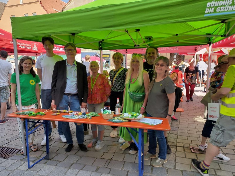 Gruppe von acht Personen steht unter einem grünen Pavillon mit dem Logo von Bündnis 90/Die Grünen auf einem belebten Straßenfest. Sie stehen hinter einem Infotisch mit Flyern, Saatgutpäckchen und kleinen Giveaways. Im Hintergrund sind weitere Stände anderer Organisationen wie der SPD und IG Metall zu sehen. Die Atmosphäre ist sonnig und freundlich.