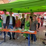 Gruppe von acht Personen steht unter einem grünen Pavillon mit dem Logo von Bündnis 90/Die Grünen auf einem belebten Straßenfest. Sie stehen hinter einem Infotisch mit Flyern, Saatgutpäckchen und kleinen Giveaways. Im Hintergrund sind weitere Stände anderer Organisationen wie der SPD und IG Metall zu sehen. Die Atmosphäre ist sonnig und freundlich.