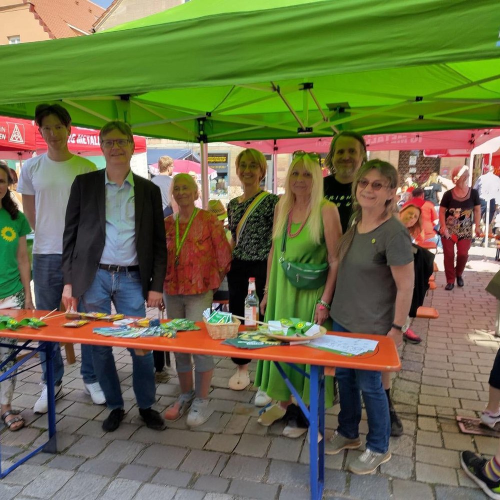 Gruppe von acht Personen steht unter einem grünen Pavillon mit dem Logo von Bündnis 90/Die Grünen auf einem belebten Straßenfest. Sie stehen hinter einem Infotisch mit Flyern, Saatgutpäckchen und kleinen Giveaways. Im Hintergrund sind weitere Stände anderer Organisationen wie der SPD und IG Metall zu sehen. Die Atmosphäre ist sonnig und freundlich.