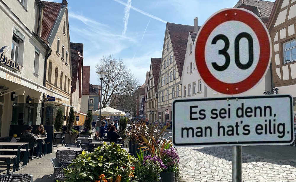 Ein belebtes Straßencafé in einer Altstadt, mit Gästen, die das sonnige Wetter genießen. Im Vordergrund ein Verkehrsschild, das ein Tempolimit von 30 km/h anzeigt, ergänzt durch ein humorvolles Zusatzschild mit der Aufschrift 'Es sei denn, man hat's eilig'. Die Szene fängt die gelassene Atmosphäre der Fußgängerzone und das Zusammenspiel von urbanem Leben und Verkehrsberuhigung ein.