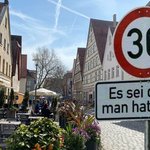 Ein belebtes Straßencafé in einer Altstadt, mit Gästen, die das sonnige Wetter genießen. Im Vordergrund ein Verkehrsschild, das ein Tempolimit von 30 km/h anzeigt, ergänzt durch ein humorvolles Zusatzschild mit der Aufschrift 'Es sei denn, man hat's eilig'. Die Szene fängt die gelassene Atmosphäre der Fußgängerzone und das Zusammenspiel von urbanem Leben und Verkehrsberuhigung ein.